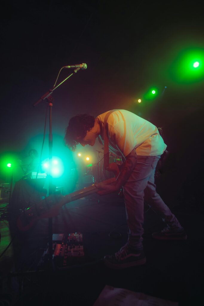 A lively musician plays guitar on stage with vibrant lights, delivering an energetic concert performance.